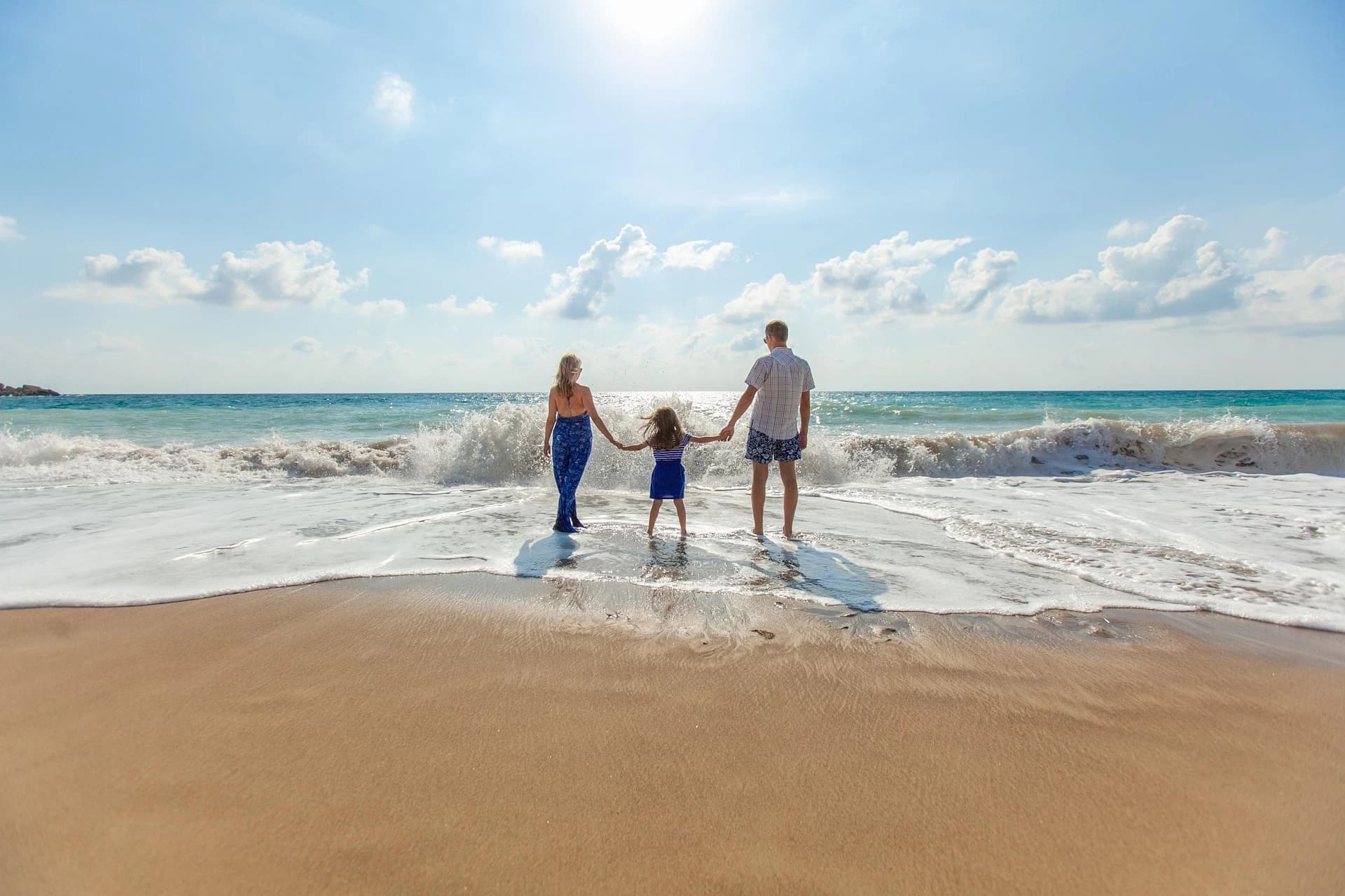Des parents et leur enfant marchent main dans la main face aux vagues sur une plage ensoleillée.