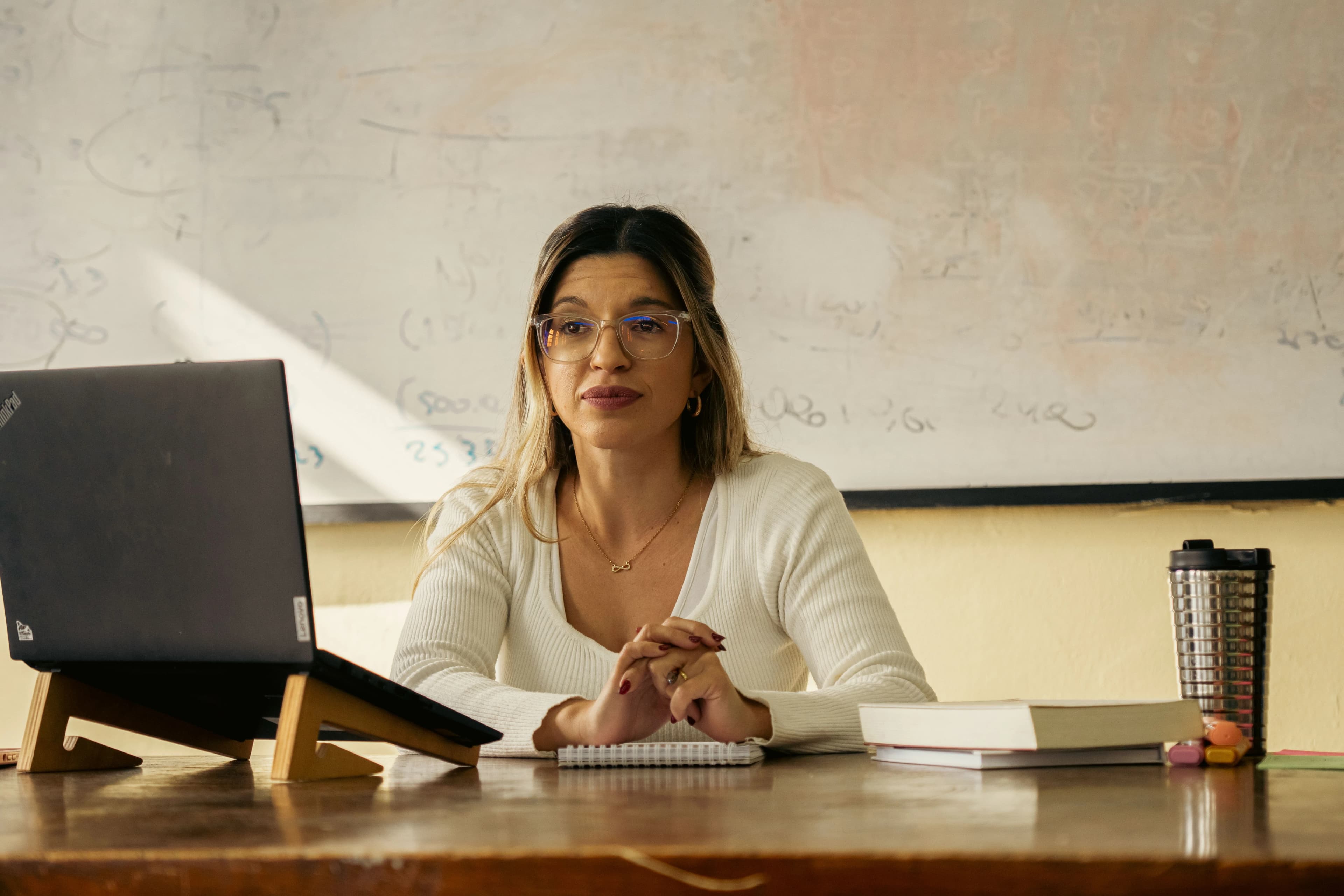 Une enseignante assise à son bureau devant un tableau blanc, ordinateur portable ouvert.