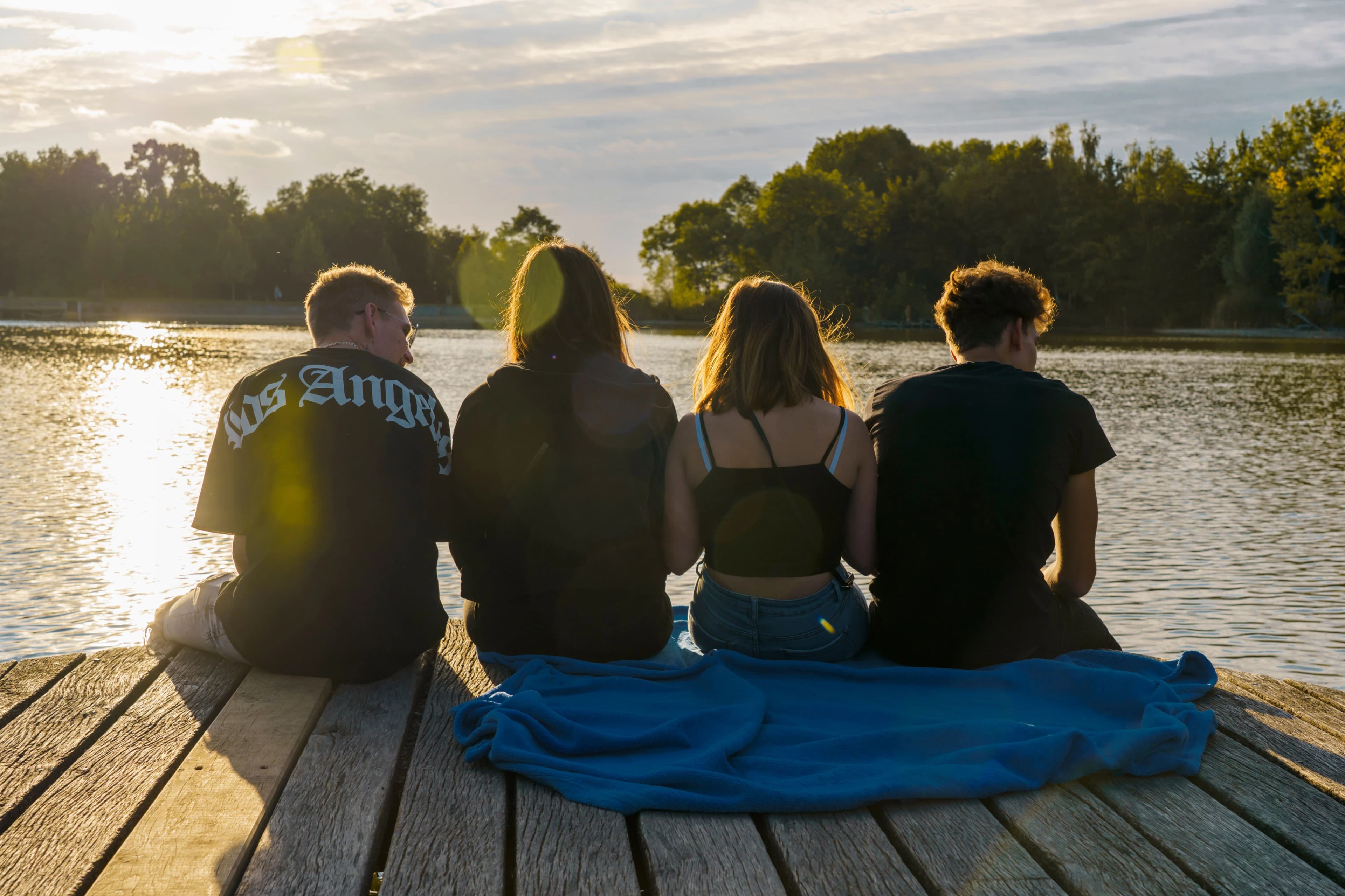 Quatre adolescents assis de dos sur un ponton au bord de l’eau au coucher du soleil, face à l’horizon.