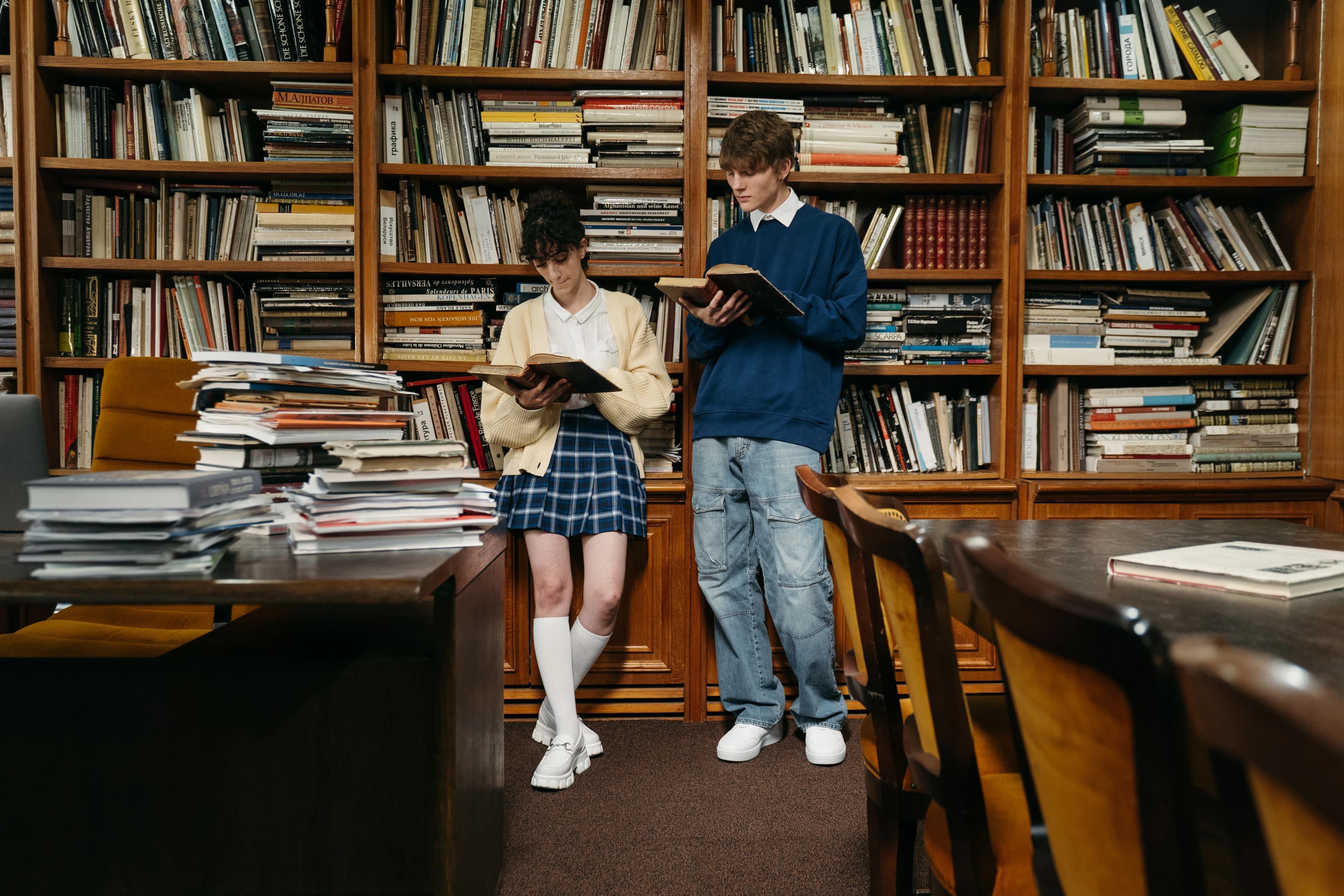 Deux lycéens lisent des livres debout devant les rayonnages d’une bibliothèque.