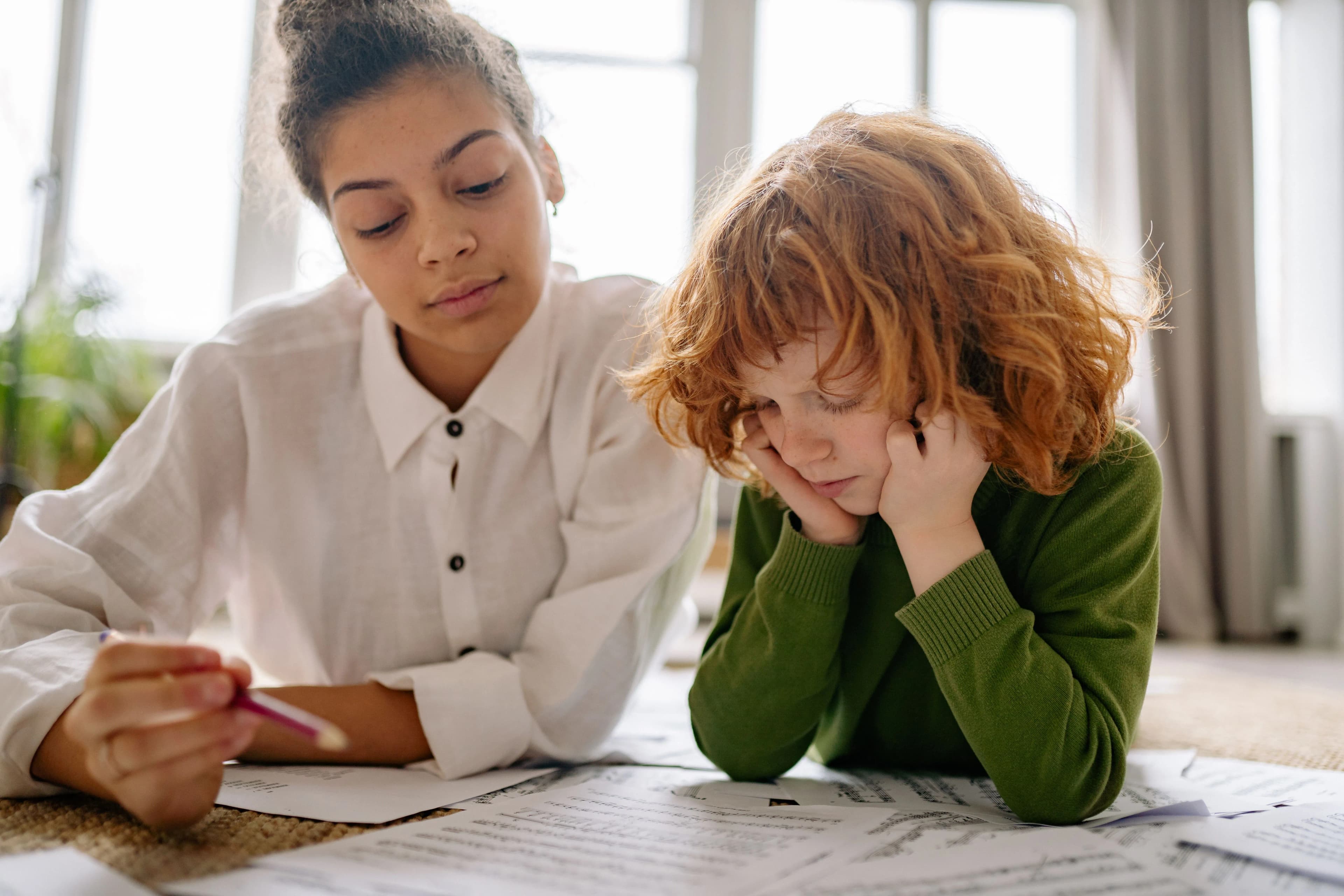 Une jeune femme accompagne un enfant pensif penché sur ses feuilles de travail.