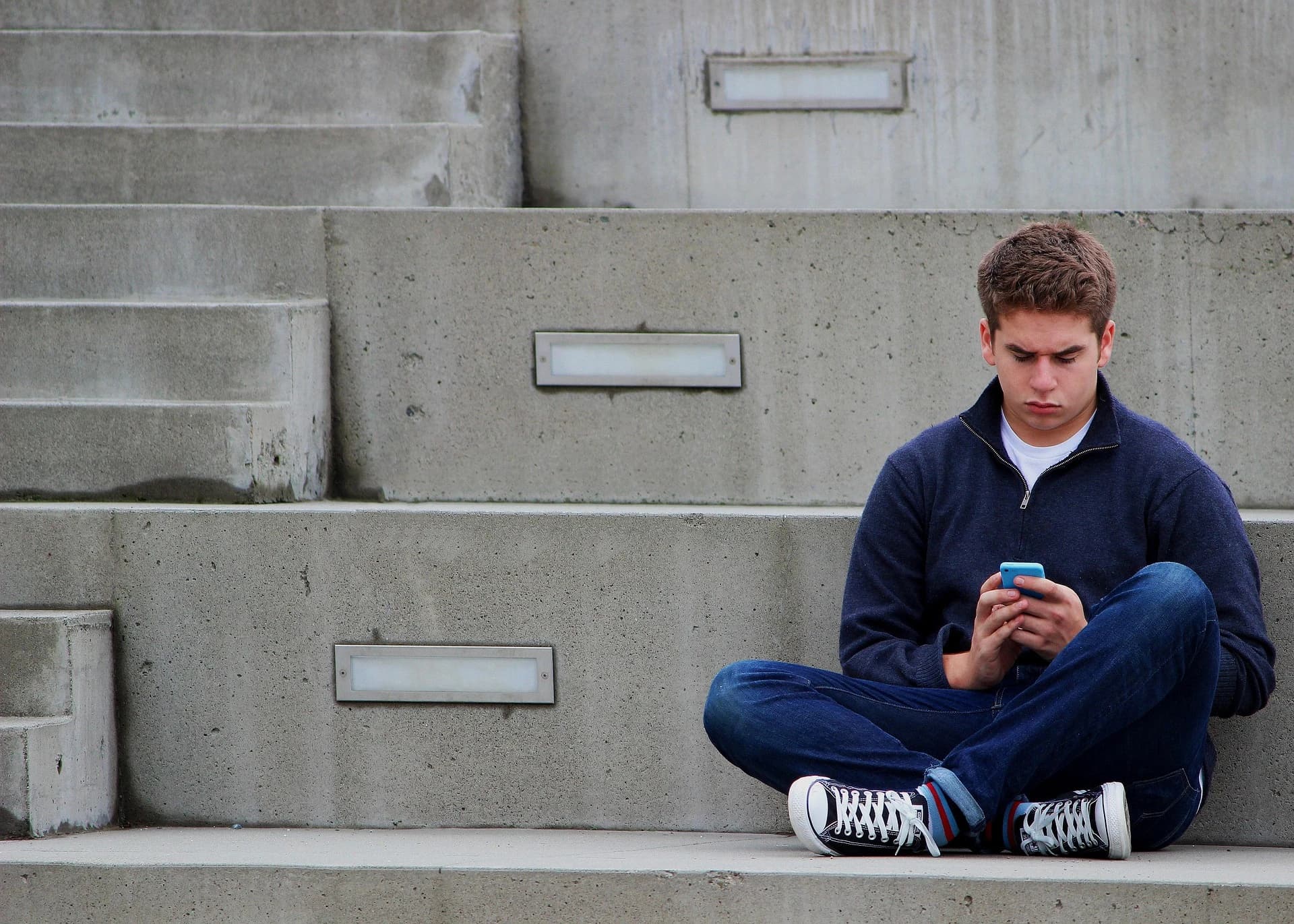 Un lycéen assis sur des marches en béton consulte attentivement son smartphone.
