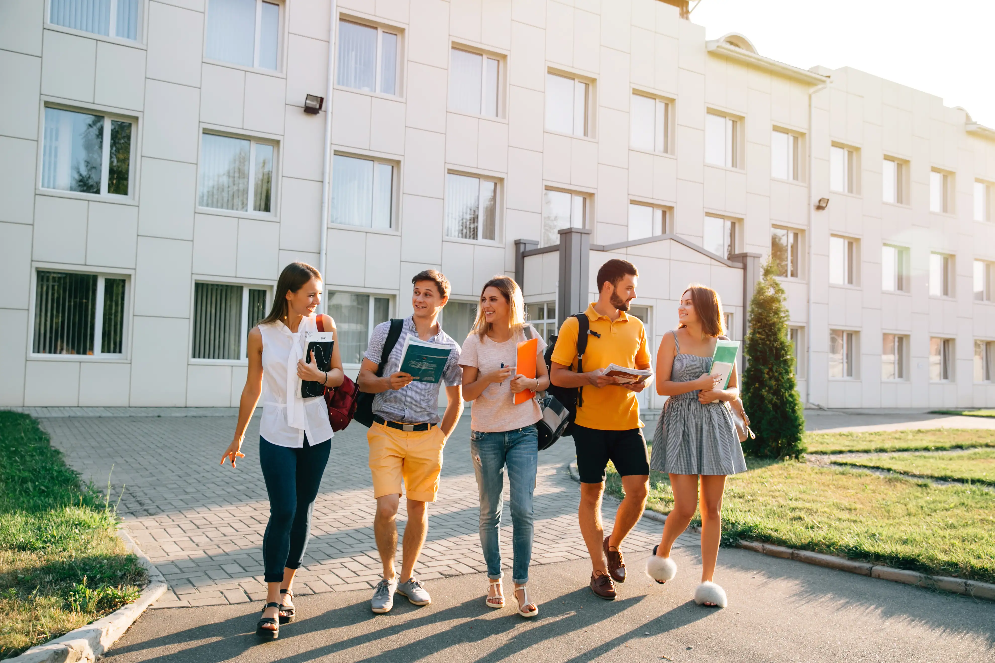 Des étudiants discutent en marchant devant un bâtiment moderne, dossiers et cahiers à la main.